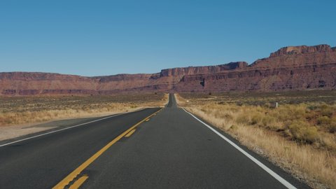 Driving on an empty road going into the distance to the red mountain rocks and hilly landscape in the arid desert. The highway with black asphalt and orange markings. Slow motion, sunny day