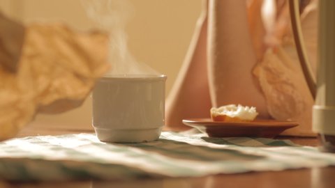 Woman sitting alone on a table holding a white porcelain cup with steaming coffee or tea beside a brown plate with a piece of bread on it