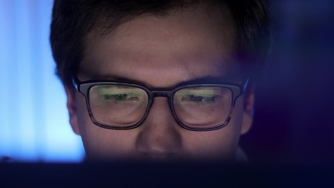 Man in eyeglasses late at night scrolling in front of laptop profile shot. Coder, programmer or developer using laptop in dark. Close up of glasses with reflection of computer screen.