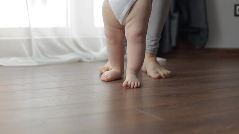 The first steps. A small child learns to walk holding his mother's hands. Caucasian child takes his first steps on the wooden heated warm floor