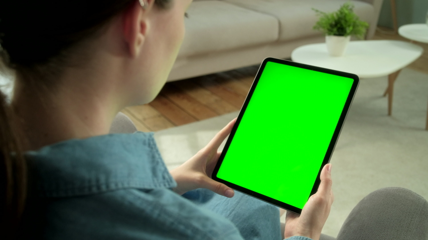 View From the Shoulder of Woman Using Hand Gestures on Green Mock-up Screen Digital Tablet Computer in Vertical Mode Sitting on a Chair. In the Background Cozy Living Room. Tapping on Center Screen | Shutterstock HD Video #1054873388