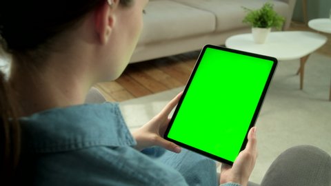 View From the Shoulder of Woman Using Hand Gestures on Green Mock-up Screen Digital Tablet Computer in Vertical Mode Sitting on a Chair. In the Background Cozy Living Room. Tapping on Center Screen