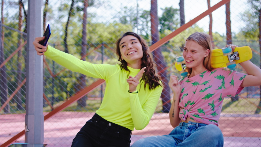 In a urban skate park two charismatic with a large smile ladies take some selfies with the smartphone they posing in front of the camera take some pictures for memories 
