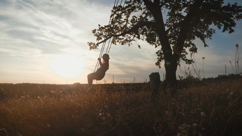 Happy child girl on swing at golden summer sunset. Silhouette of a young teenager girl swinging on the lone tree at nature, lifestyle. The concept of childhood dreams.