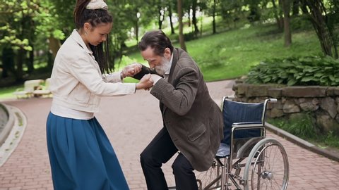Likable lucky bearded 70-aged man trying to get up from wheelchair and appealing satisfied cheerful young girl with long dreadlocks helping him,holding by hands,slow motion
