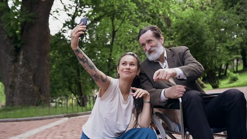 Front view of good-looking satisfied joyful 25-aged girl with tatoo on arm and long dreadlocks and cheerful esteemed mature grandfather in wheelchair with grey beard which making photos on mobile in