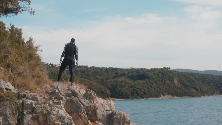 Male looking at green forested hills and blue cloudy sky. Stock. Rear view of a man in black sports suit standing on the rock by the sea or river and looking into the distance.