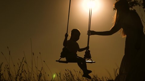 baby and mom swinging on swing in park in sun. silhouette of small healthy child on swing. daughter and mother love flying on swing on summer evening in forest. concept of happy family and childhood