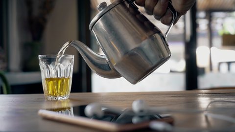 Close-up smartphone with headphones and hand with a steel traditional teapot pouring tea into a clear glass on a wooden table in a street cafe