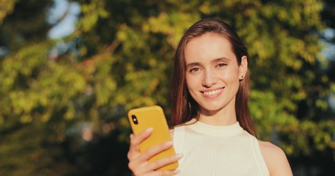 Close up view of beautiful brunnette woman looking to camera and smiling while using smartphone . Happy girl scrolling phone screen while standing outdoors. Blurred park background.