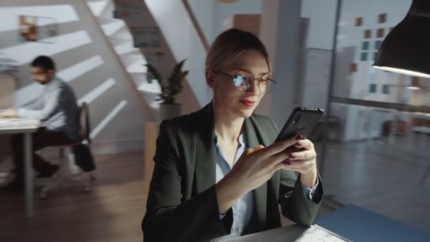 360-degree dolly shot of cheerful blonde businesswoman in formalwear and glasses sitting at desk and texting on smartphone while working in the office