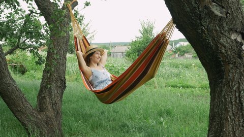 Young cute brunette girl resting on hammock, unwinds on hammock in countryside. Caucasian woman in hat and dress relaxing and raising hands while lying on hammock outdoors