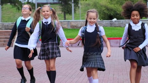 adorable little schoolgirls in uniform with skirts run along school yard against flowerbeds in warm summer slow motion
