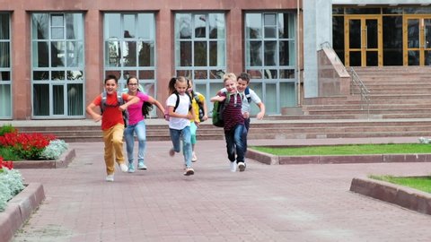 group of joyful little classmates in casual clothes with bags run along school yard with flowerbeds on warm day slow motion