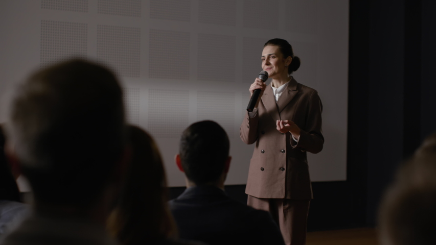Speech of female employee at economy summit. Independent speaker talks at scene for business men in crowded conference hall. Woman explains motivation to group of people. Teamwork meet at modern event Royalty-Free Stock Footage #1054902014