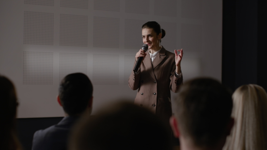 Speech of female employee at economy summit. Independent speaker talks at scene for business men in crowded conference hall. Woman explains motivation to group of people. Teamwork meet at modern event Royalty-Free Stock Footage #1054902014