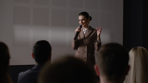 Speech of female employee at economy summit. Independent speaker talks at scene for business men in crowded conference hall. Woman explains motivation to group of people. Teamwork meet at modern event