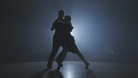 Silhouette of dancers in smoky ballroom. Young couple dance tango in dark room with smoke and spotlights in slow motion. Wide angle in 4K, UHD