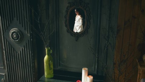 Interior dark gothic room medieval fairytale castle. Candles burning on black vintage table, red enchanted apple, bare tree branches in green vase, ghost reflection of beautiful woman in old mirror