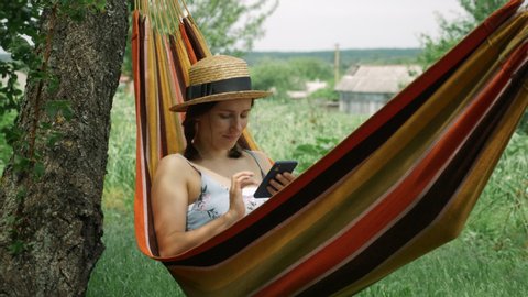 Portrait of young woman using mobile phone lying in hammock outdoors. Brunette female in hat resting in hammock with smartphone at green garden. Cute charming girl relaxing in hammock countryside
