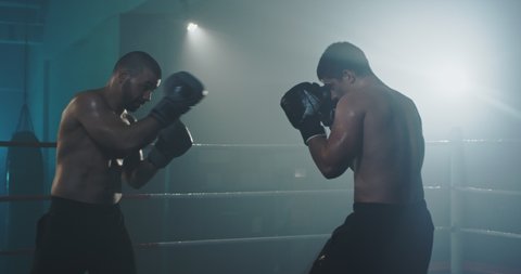 Musculars young Caucasian men fighters of box at ring. Male boxers fighting in dark. Shirtless kickboxers punching each other while training in gym.