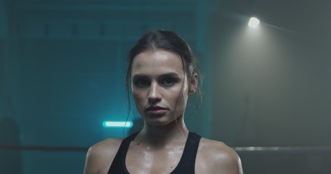 Portrait of beautiful young tired and sweaty woman boxer turning face to camera with serious look at dark ring. Close up of pretty girl sportswoman looking at cam after physical training.
