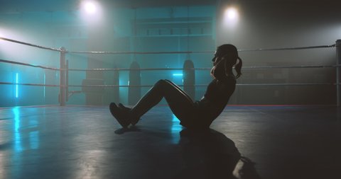 Caucasian young woman in good body shape lying on ring floor and doing abs. Flat belly resolution. Sportswoman training before boxing in darkness. Female boxer warming up and working out.