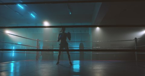 Dark female silhouette of boxer in gloves coming in the ring. Girl kickboxer training alone in darkness. Slim woman, kickbox fighter working out. Box practise concept.
