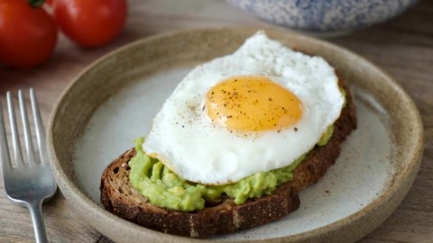 Rye bread toast with mashed avocado and sunny side up egg. Cutting egg yolk with a knife. Tasty healthy sandwich