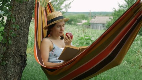 Portrait of young brunette woman in straw hat lying in hammock, reading book and eating apple. Happy brunette female resting in hammock outdoors. Relaxed girl in hammock at green garden