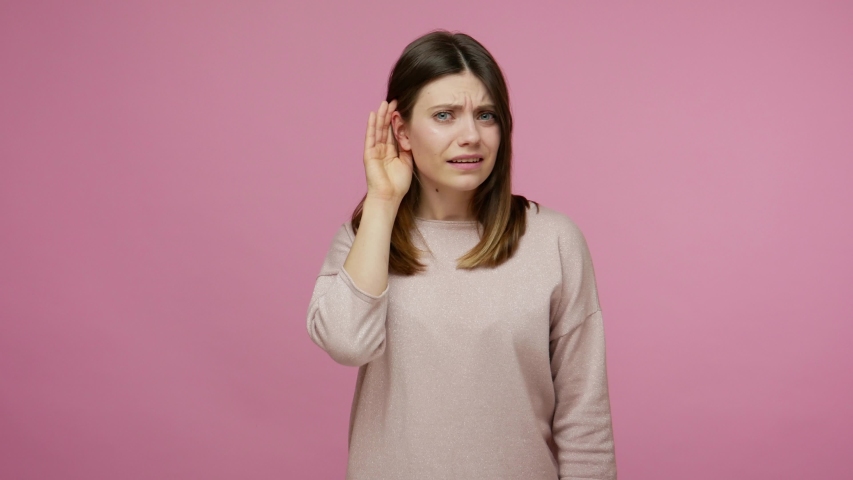 Brunette woman listening to conversation with hand near ear, expressing doubts, unsure of heard information, suffering hearing problems and deafness at young age. indoor studio shot, pink background | Shutterstock HD Video #1054912562