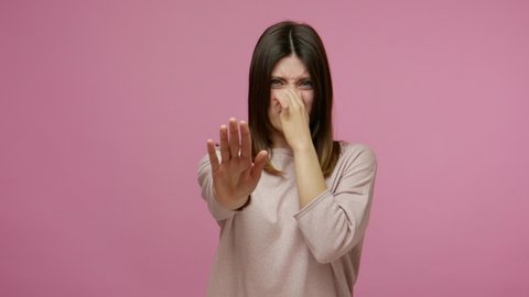 Repulsion to bad smell. Brunette girl grabbing nose with fingers, holding breath to avoid stink, awful fart gases, intolerable odor, showing stop gesture. studio shot isolated on pink background