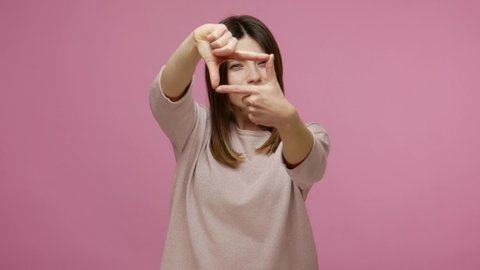 Concentrated attentive brunette woman looking through finger frame gesture, imitating photo zoom, capturing interesting moment, focusing at target. indoor studio shot isolated on pink background