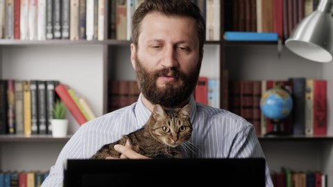 Man with cat working at computer. Focused bearded male worker freelancer businessman salesman at workplace holding cat and typing on laptop keyboard enters data. Close-up