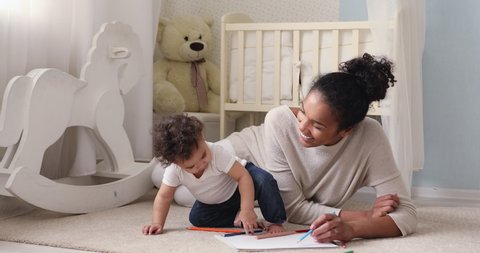 Adorable african american small toddler kid sitting on floor carpet with smiling biracial mom nanny, holding colorful pencils in hands. Loving mixed race mum teaching little infant drawing pictures.