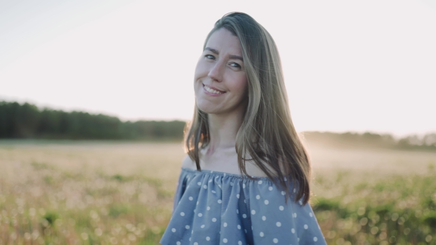 A caucasian beautiful wonan standing on the beautiful field during summer sunset and looking at the camera. The woman is dancing. The woman wears beautiful dress. Close up.