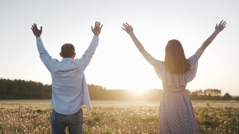 A caucasian man and woman standing on the beautiful field during summer sunset. Their hands are raised. A man and woman stand with their back to the camera. Close up.