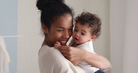 Head shot loving young african ethnicity mother holding cute funny kid daughter, kissing her cheeks. Affectionate mixed race nanny mum enjoying sweet tender moment with adorable baby son at home.
