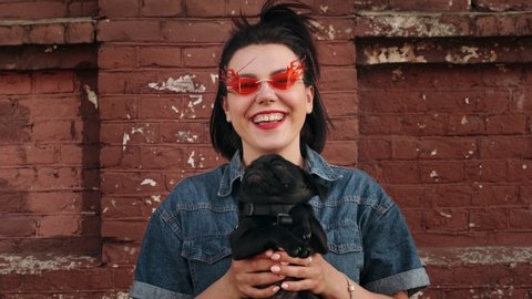 Stylish woman in fashionable red glasses laughs and shakes a funny pug puppy in her arms. Portrait student, millennial dog lover.