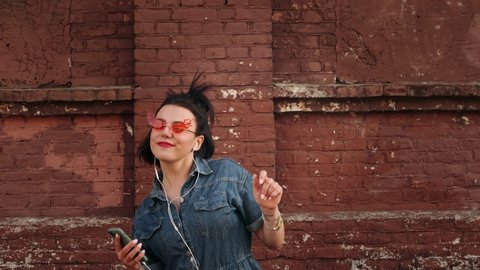 Stylish hipster woman using mobile phone touch screen and  listening to music by earphone at city urban background. Woman enjoying music, dancing in color fashion glasses.