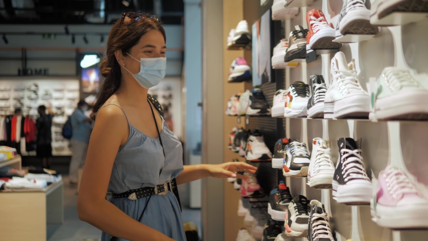 Young brunette woman girl in medical mask choosing shoes in a shoe store. Shopping mall, safety during coronavirus pandemic. Business after covid quarantine. | Shutterstock HD Video #1054918964