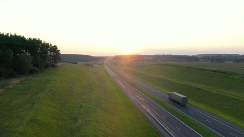 Truck driver with a cargo is driving on an intercity speed highway to the customer through a picturesque rural area with a sunset background. Delivery and logistics of goods , aerial motion forward