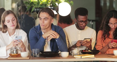 Millennial friends using their phones while spending time together in cafe. Young guy looking bored while sitting between gadget addicted multi ethnic people with smartphones
