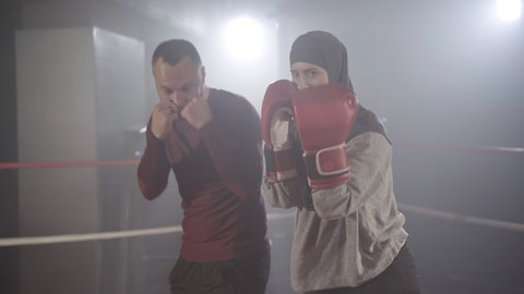 Coach correcting boxing stance of young beautiful muslim woman in hijab training in haze in backlight. Portrait of confident female boxer exercising with personal trainer in lense flare and fog.