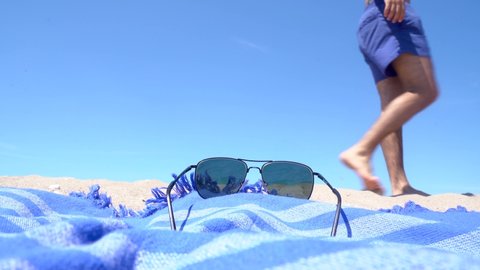 Person enjoying the summer beach holiday wearing a blue swimsuit, blue beach towel and sunglasses on the sandy beach on a sunny day.
