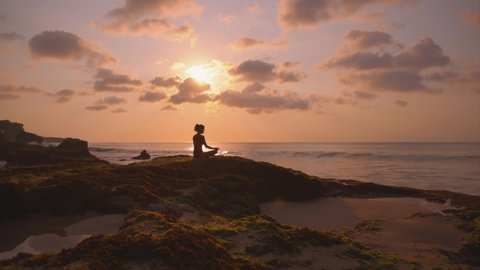 Silhouette of young woman relaxing and enjoy beauty sunset on the ocean coast. Girl doing yoga on the beach. Person meditate outdoor and enjoy orange sunlight. Concept of fitness, health, relax, yoga.