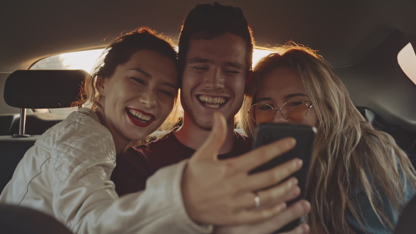 Beautiful young girls taking selfie with their friend on phone or smartphone in back seat of moving car while roadtrip. Cheerful friends having good time together. Concept of joy and lifestyle Royalty-Free Stock Footage #1054928903