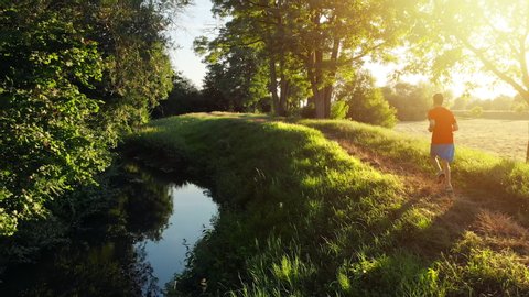 Dynamic fitness footage of a runner on a narrow path, running in nature along a stream at sunset, being followed by the camera 