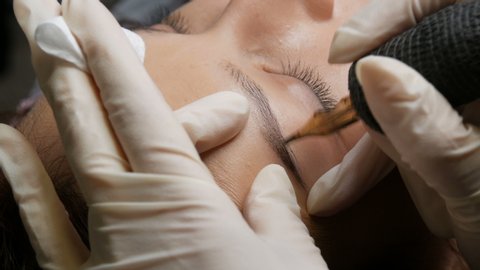 Beautician in black gloves makes permanent makeup correction of the shape of the eyebrows to a young beautiful girl. Microblading, eyebrow tattoo in a beauty salon.