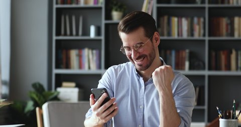 Excited young businessman in eyeglasses reading message with good news on smartphone. Happy euphoric male user looking at cellphone screen, celebrating online win, mobile victory success concept.